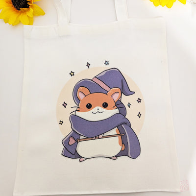 Cute kawaii wizard hamster tote bag with pastel fantasy artwork featuring a magical hamster holding a wand and spell book. Reusable 14 x 16 inch canvas polyester shoulder bag for shopping, school, and everyday use.
