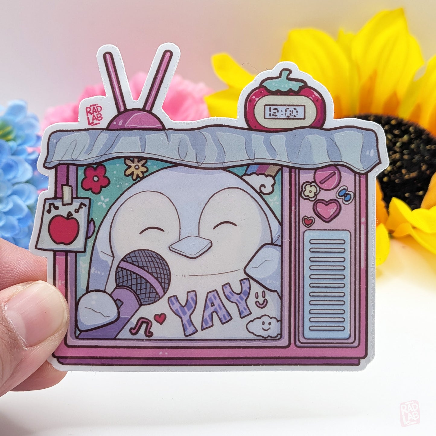 Cartoon sticker of a penguin with a microphone and 'YAY' text, held against a colorful background.